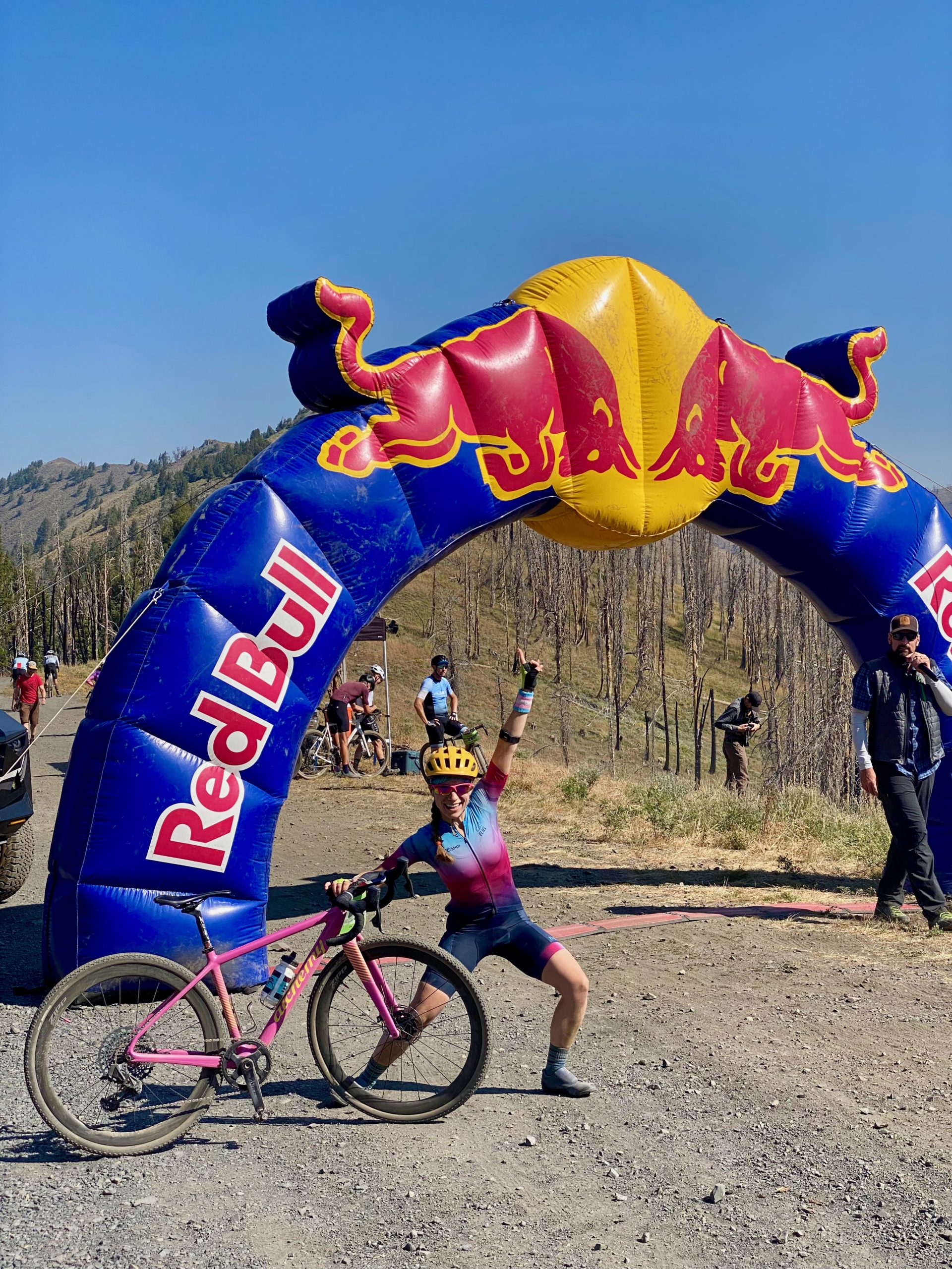 Woman in bike kit poses triumphantly with her bike under a large Red Bull banner