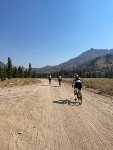 Three cyclists ride on a gravel road with mountains on the left side