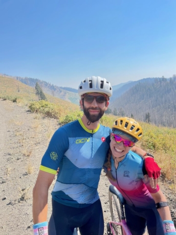 Man and woman pose together in bike jerseys with mountains in the background