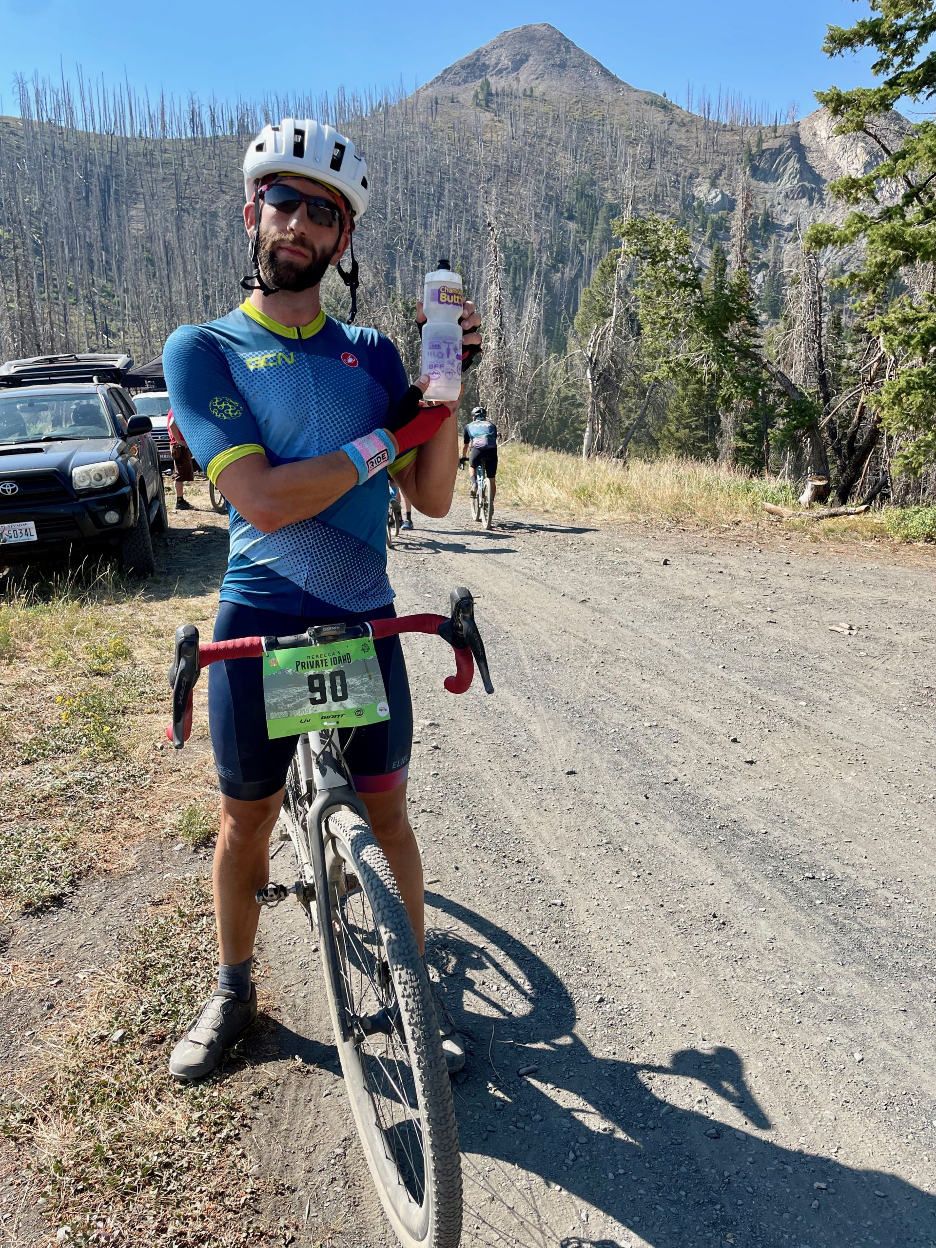 Man standing over bike on dirt road holds a water bottle from Chamois Butt'r