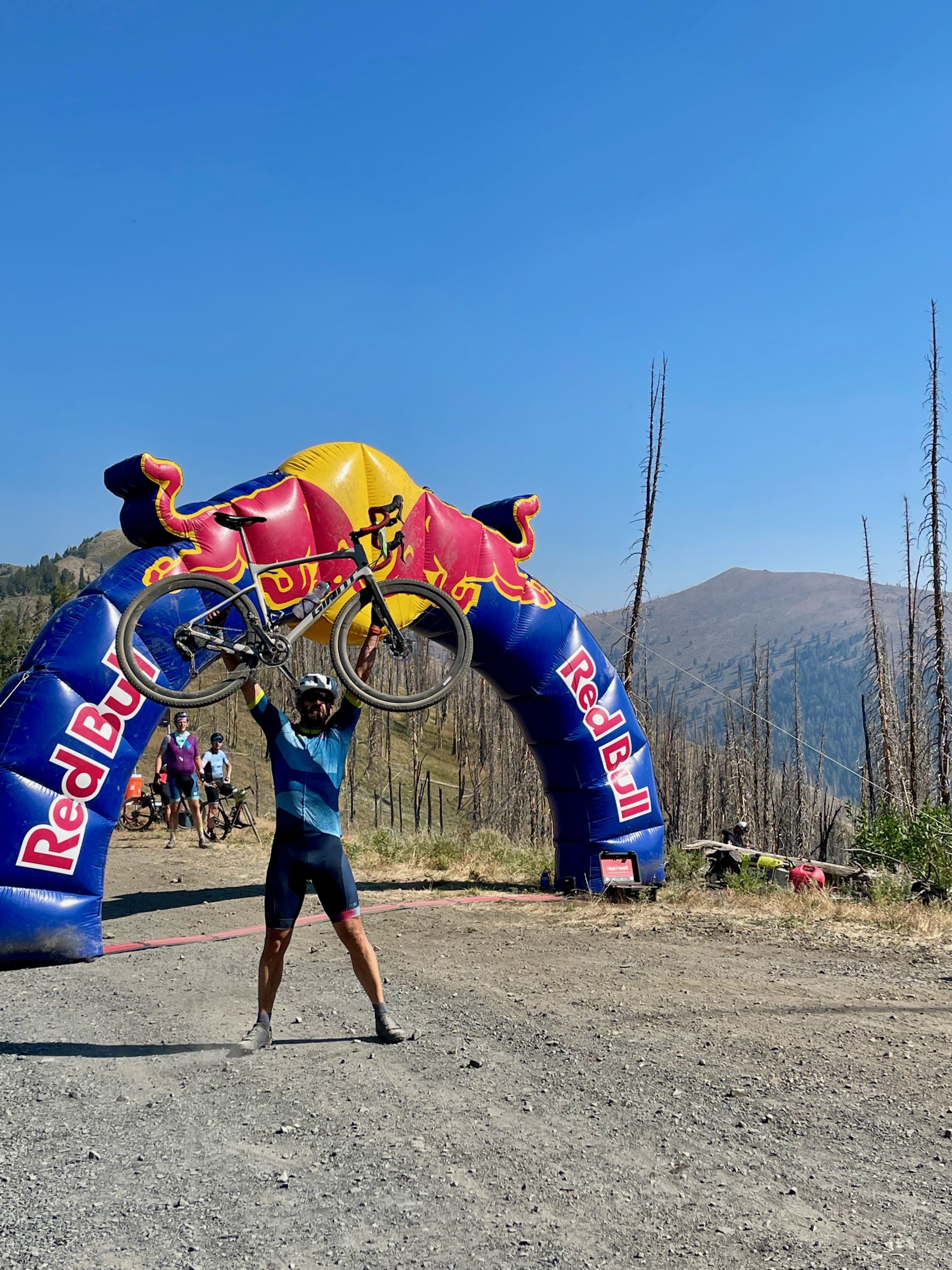 A man stands holding his bicycle over his head in front of a large Red Bull banner on a dirt road