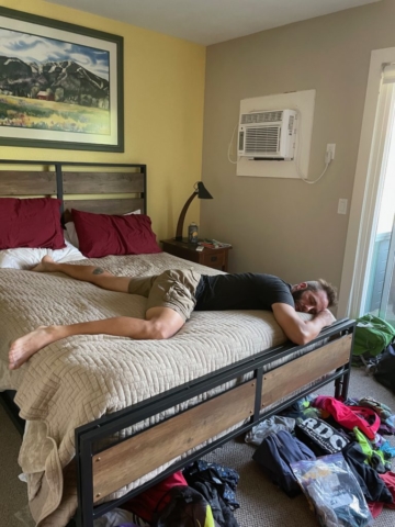 Man sprawled across bed under a wall AC unit in shorts and t-shirt