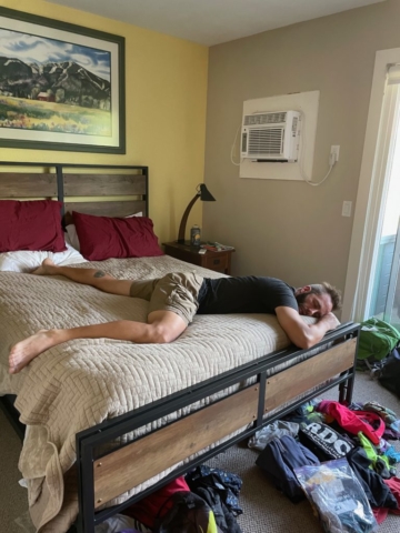 Man sprawled across bed under a wall AC unit in shorts and t-shirt