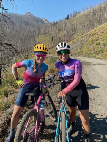 Two woman in cycling kit and helmets stand next to their bikes on a dirt road with mountains in the background
