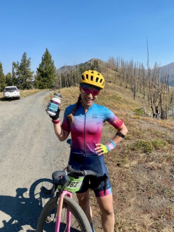 Woman stands over bike holding a BaseCamp water bottle in BaseCamp pink and blue cycling kit and a yellow helmet
