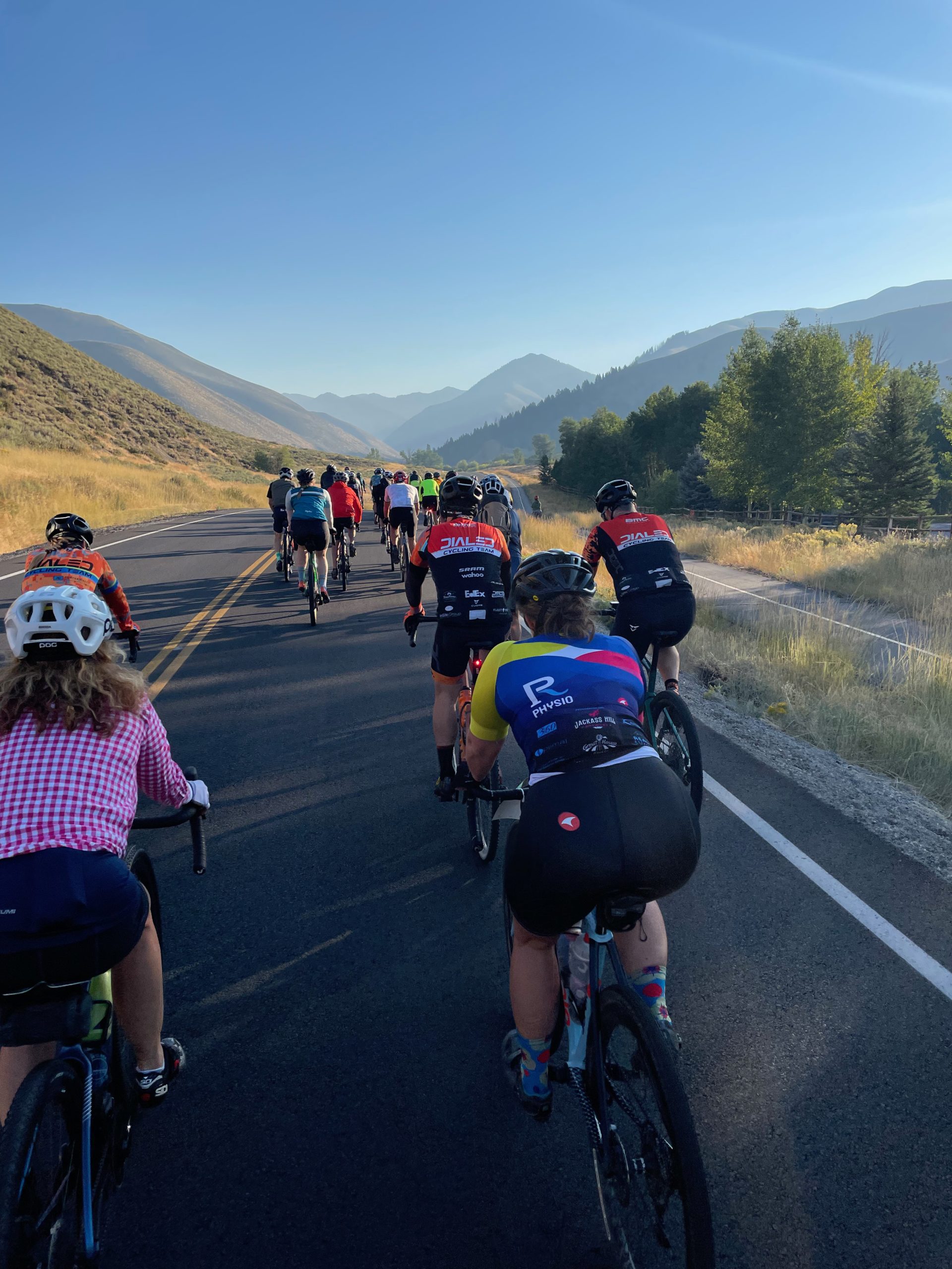 A group of over a dozen cyclists ride on a paved road in early morning with mountains in the distance